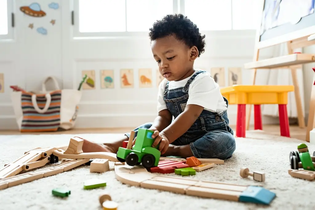 Toddler in denim dungarees sitting on a cream rug, rolling a green wooden train along a wooden track