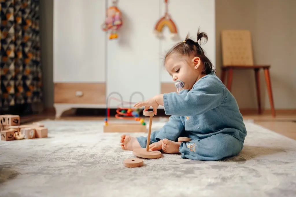 Toddler in a blue knitted outfit sitting on a soft rug, threading a wooden ring onto a stacking toy in a cosy playroom